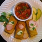 Chicken flautas topped with sliced radishes, avocado cream sauce, cilantro and side of homemade salsa on a white ceramic plate.