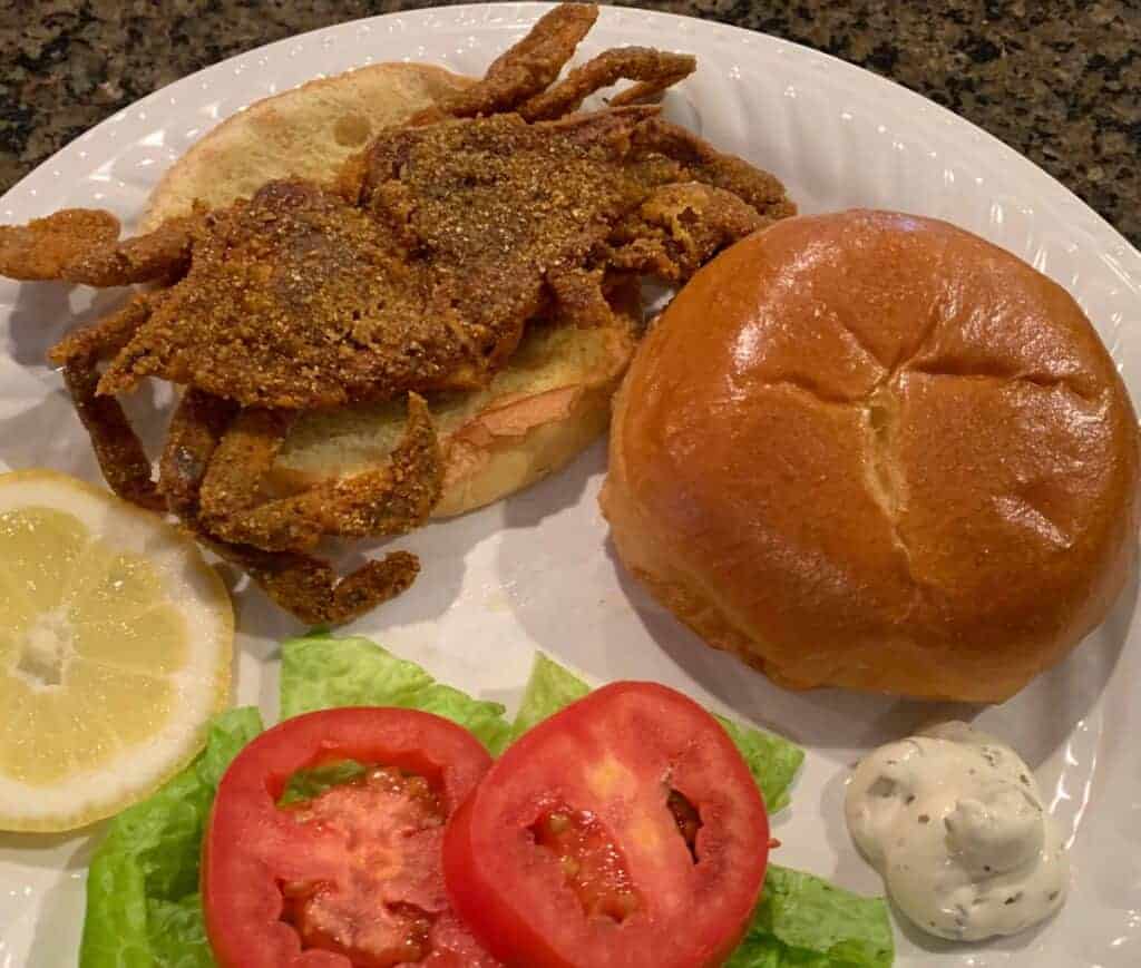 Soft shell crab sandwich on a brioche bun with lettuce and tomato with slice of lemon and tartar sauce served on a white plate
