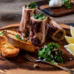 Roasted Marrow Bones with Parsley Salad, Crusted Bread, Lemon Slices, with Coarse Salt, Garlic Bulb and Red Wine Glass.