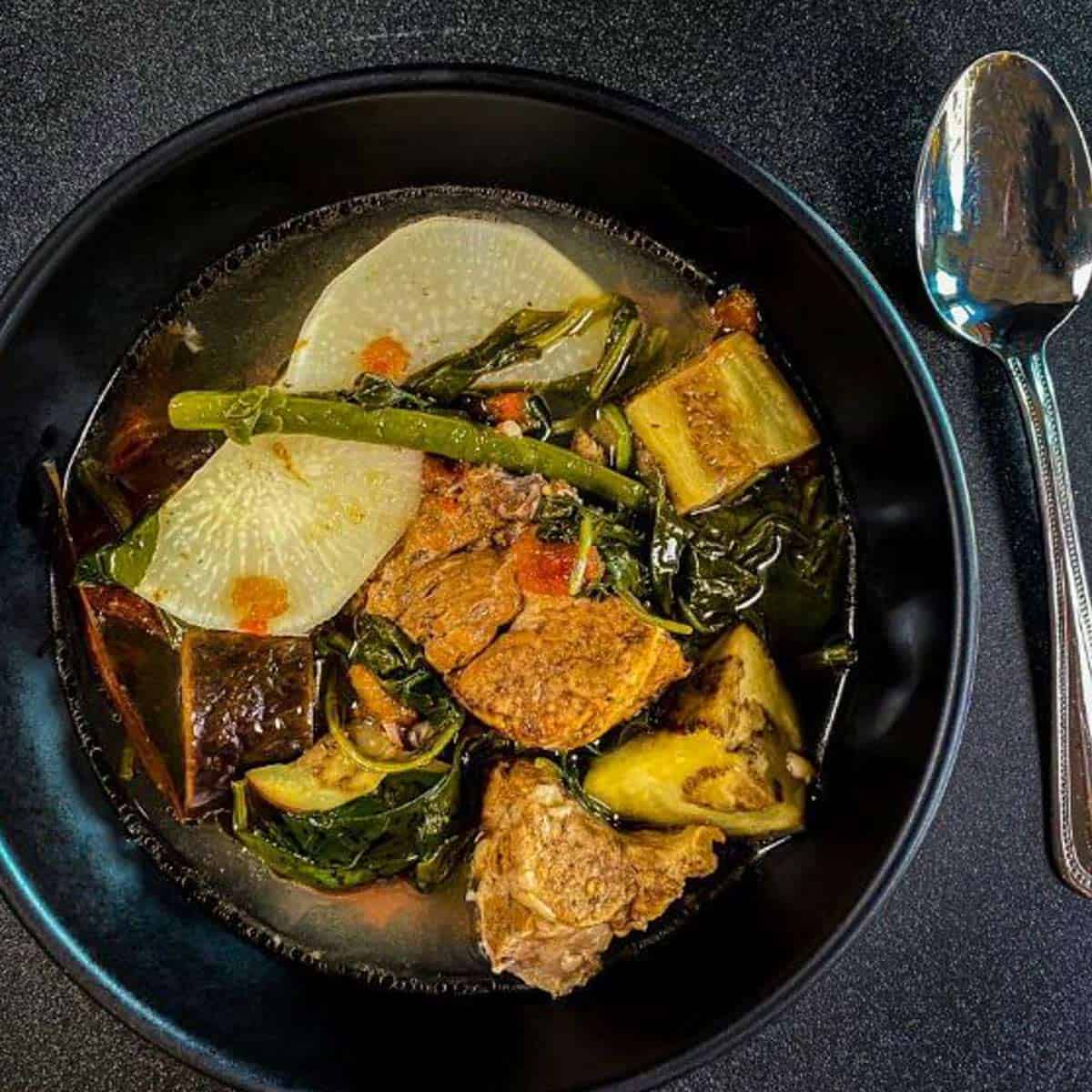 Sinigang Pork in black bowl with rutabaga, eggplant, spinach, green beans and tomatoes