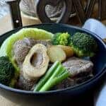 beef shank with broccoli, cabbage, green onion and potatoes in a black bowl, with spoon and napkin on the side.