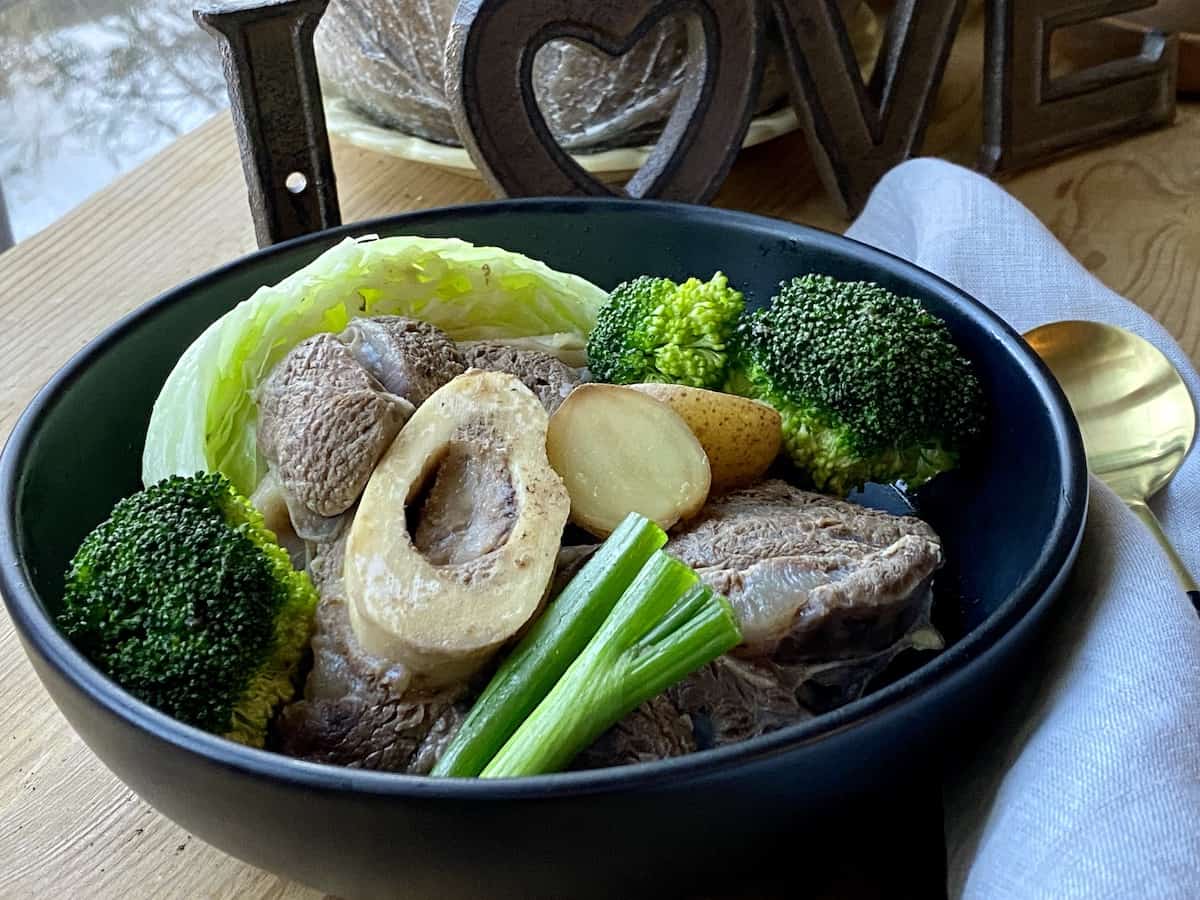 Beef shanks with broccoli, cabbage and green onion in a black bowl with a soup spoon on the side.