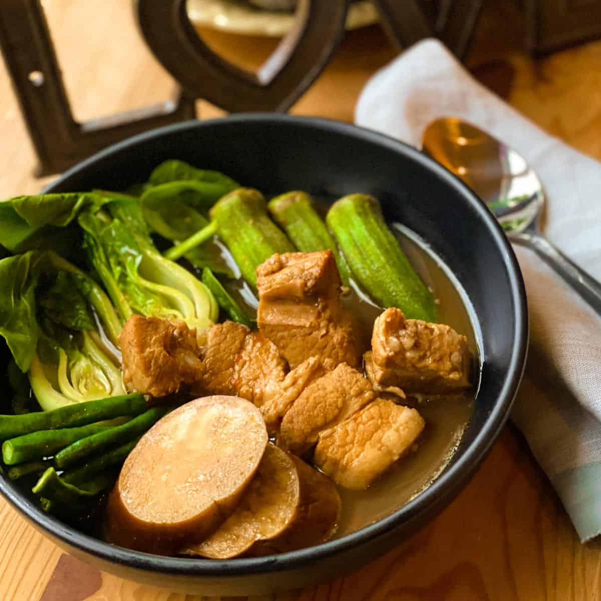 Pork sinigang sour soup with sliced eggplant, string beans, baby bokchoy, okra with tamarind sauce in a black bowl with soup and napkin on the side