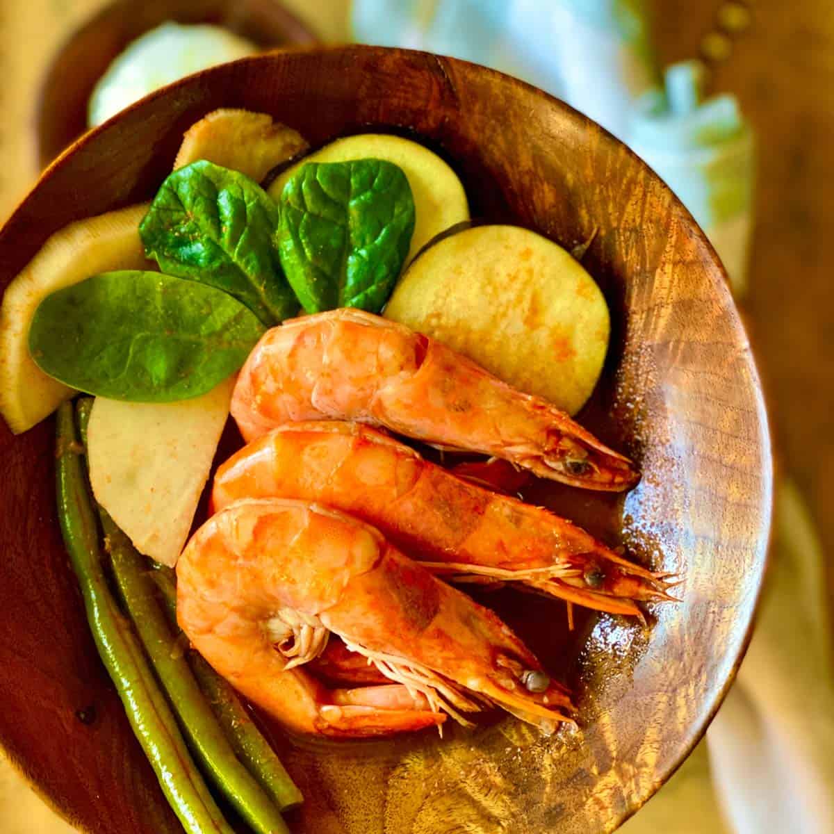 Two bowls of shrimps with eggplant, radish, greenbeans, and spinach on top of a table with napkin and a bowl of rice next to them.