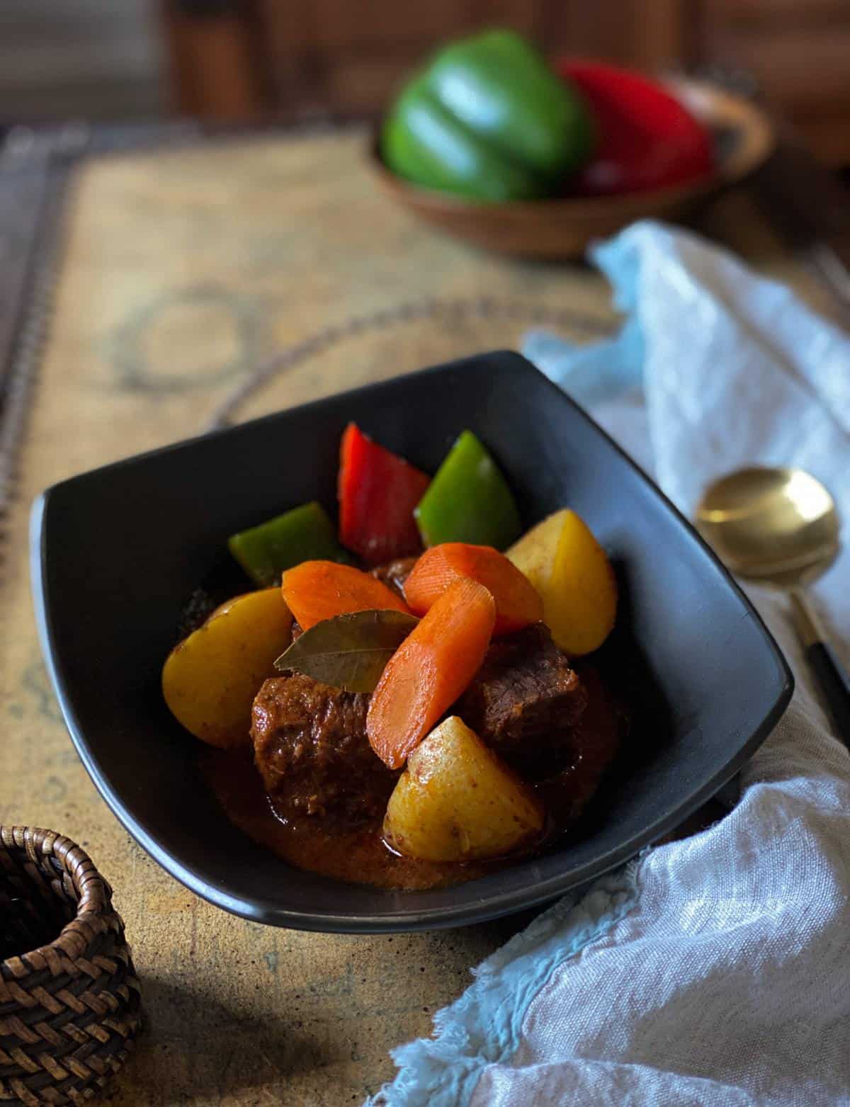 Beef with carrots, potatoes, and red and green bell peppers in a black dish on a table with napkin and spoon on the side.