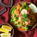 Pancit Canton Noodles with shrimp and vegetables on a wooden plate with lemon wedges on the side.