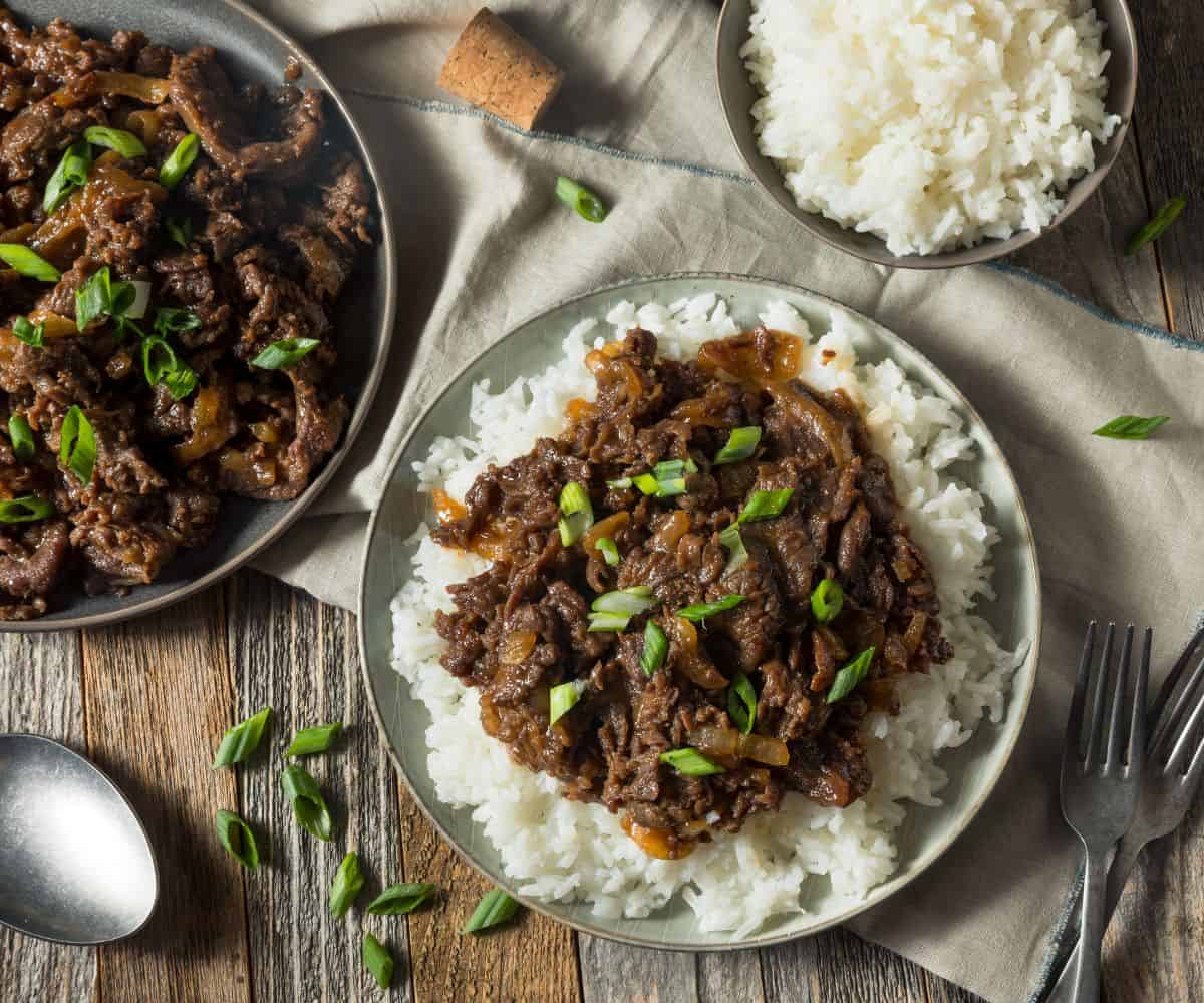 Marinated Beef with scallions on a bed of white rice on a plate with forks on the side.