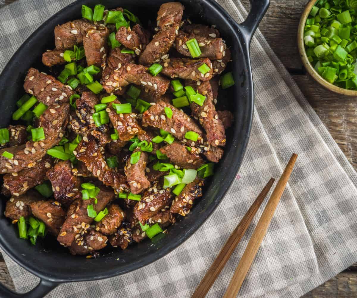 Marinated Beef with sesame seeds and scallion in a black skillet with chopsticks on the side.