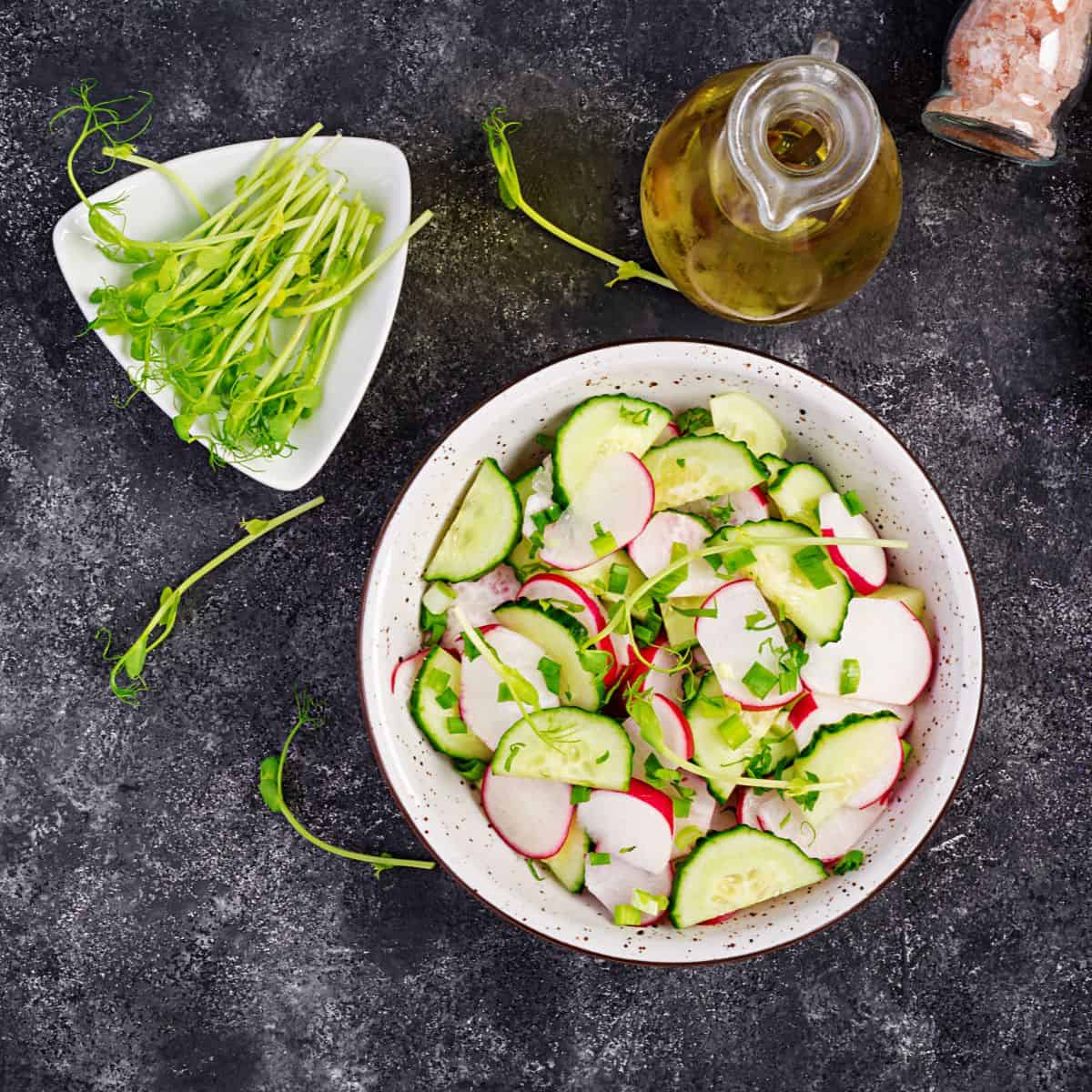 Sliced cucumbers and radishes in a white bowl on a black counter top with olive oil container on the side.