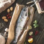 Whole salmon on a brown paper bag with herbs and spices on the side of a wooden board.