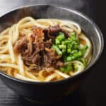 Beef udon with sliced green onions in a black bowl.