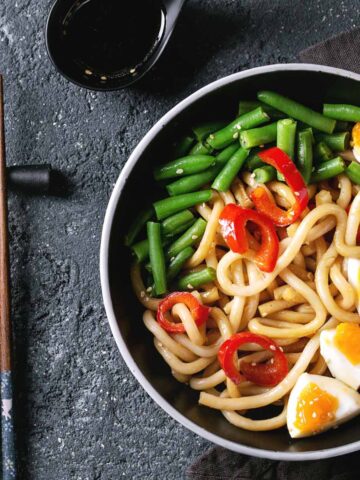 Udon with sliced green beans and hard boiled egg in a black bowl with chopstick on the side.