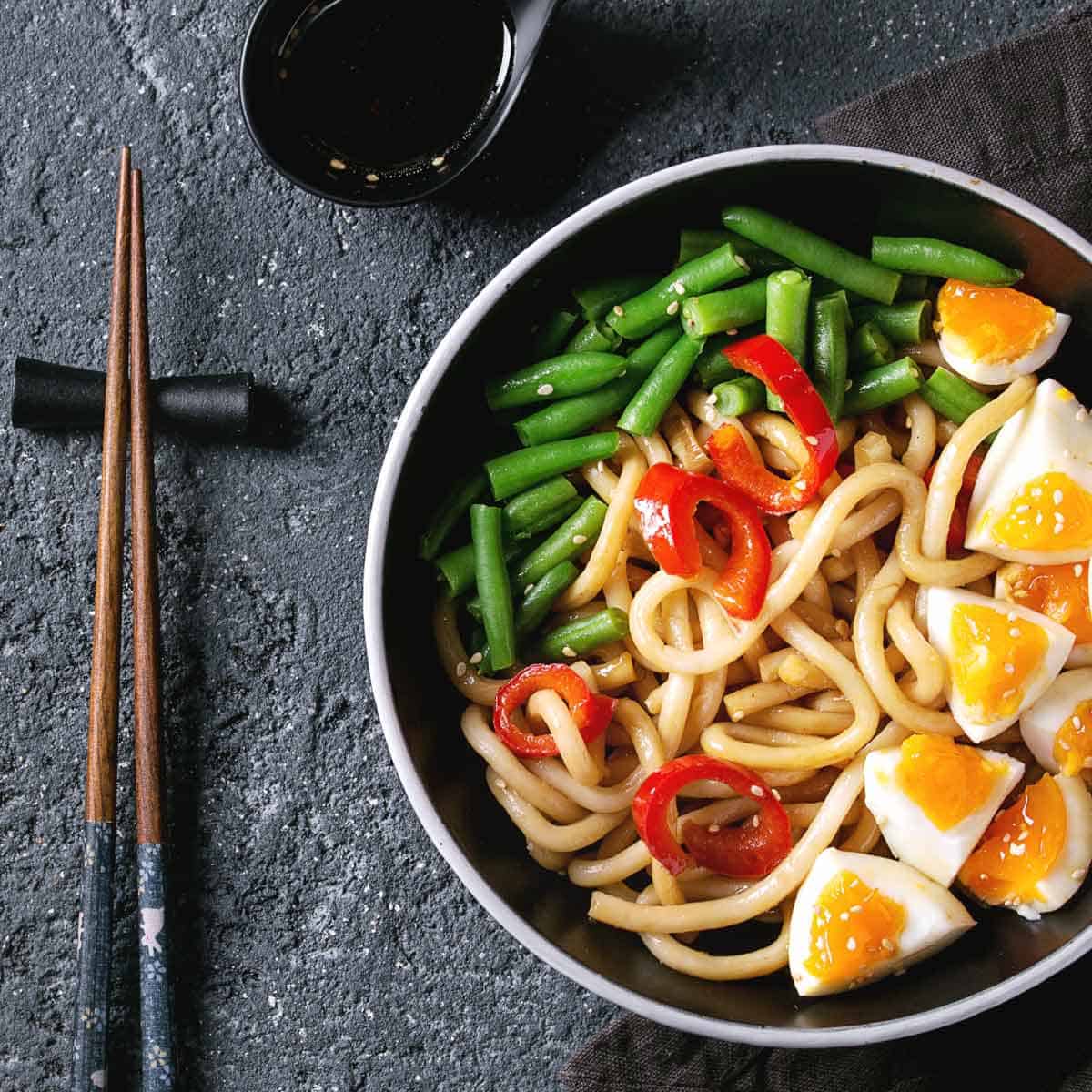 Udon with sliced green beans and hard boiled egg in a black bowl with chopstick on the side.